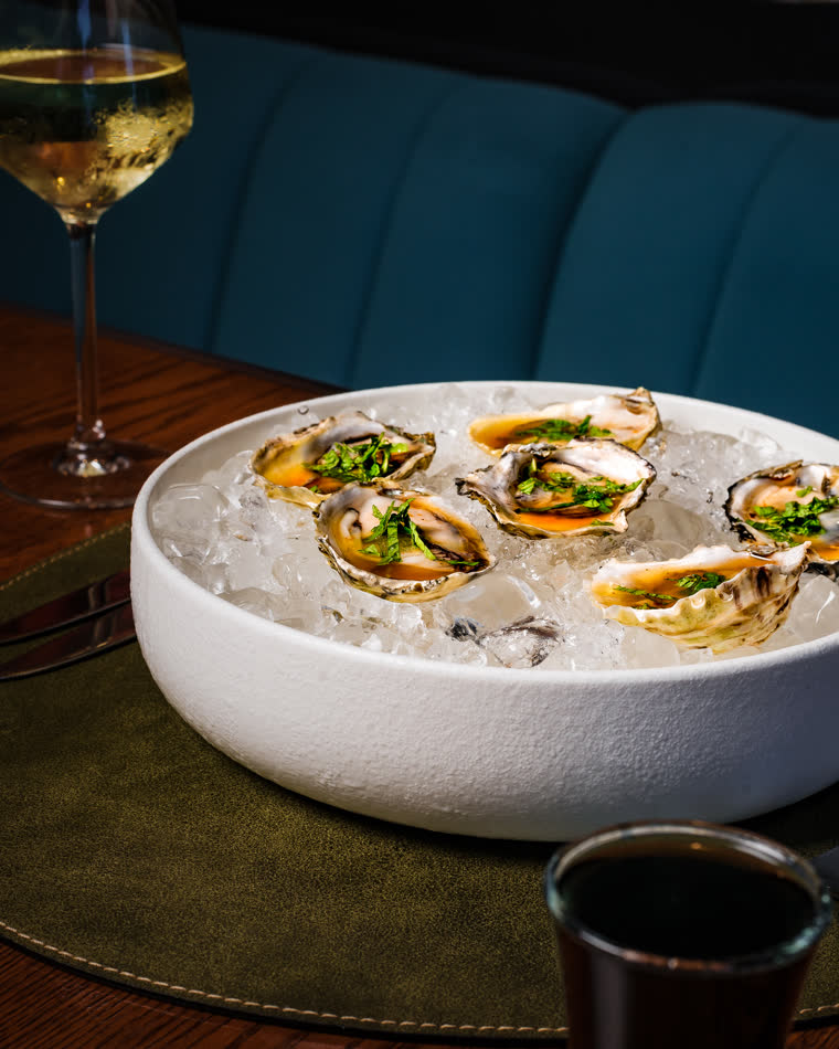 A bowl of oysters on ice beside a glass of white wine in the dining room